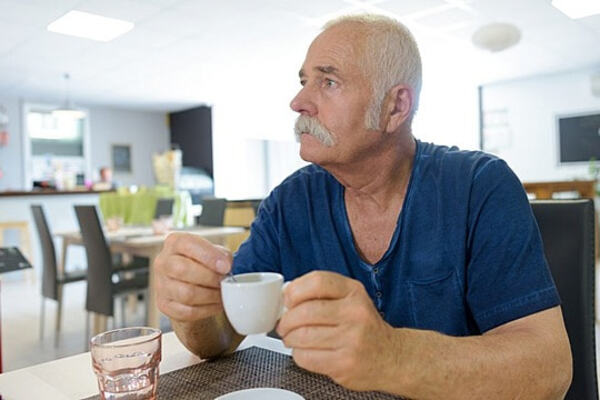 Възрастен човек седи до прозорец вечер с чаша топла напитка и мека светлина